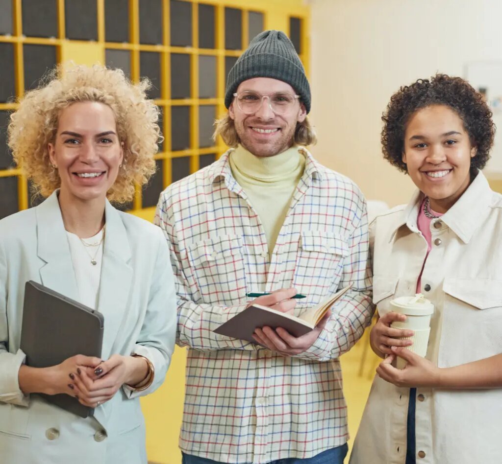Three smiling creative marketing professionals standing together in a modern office in front of a yellow accent wall. The woman on the left holds a tablet, the man in the center wears a grey beanie and glasses while holding an open notebook, and the woman on the right holds a reusable coffee cup.