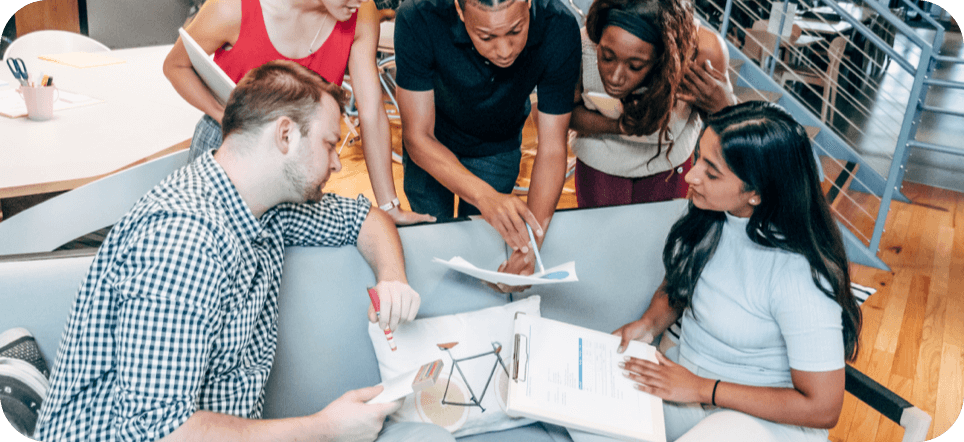 A diverse group of creative professionals gathered closely around a table collaborating on a business strategy. One team member points with a pen to a printed chart, while others look on intently with clipboards and notes in a modern office space.
