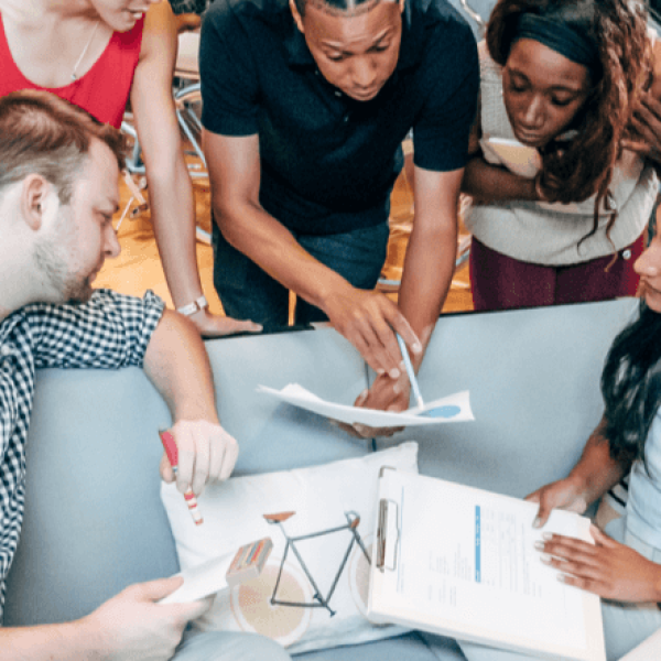 A diverse group of creative professionals gathered closely around a table collaborating on a business strategy. One team member points with a pen to a printed chart, while others look on intently with clipboards and notes in a modern office space.